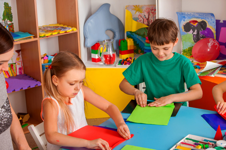 School children with scissors in kids hands cutting paper with teacher in class room. Development and social lerning. Childrens project in kindergarten together. Origami competitions.の写真素材