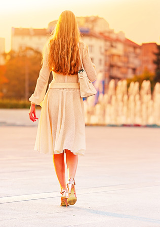 Fashion woman in autumn spring dress on city street and car. Back of female style of feminine fashionable girl model wallking hight heel fall outdoor. Color tone on shiny fountains sunlight background.の写真素材