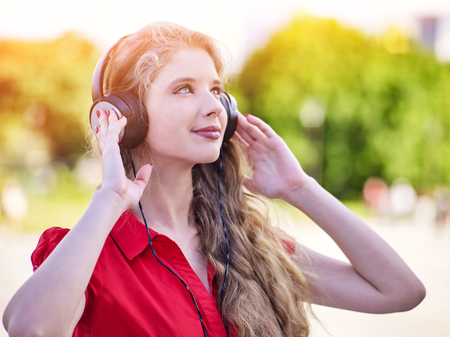 Girl in headphones and red shirt listens to mp3 music in city park. Teen portrait backlight sun flare.の写真素材