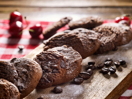 Oatmeal chocolate cookies with coffee grains and cherry, powdered sugar on kitchen cutting board with checkered fabric on wooden table in village style for picnic. Sun flare in morning sunrays.の写真素材