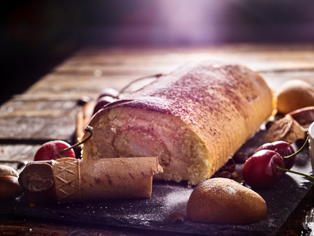 Sand cookies heart shape and chocolate rolled cake with cherry on kitchen on wooden table in countrylike style. Sun flare of sunlight and sunrays at morning.の写真素材