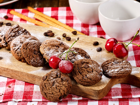 Oatmeal chocolate cookies with coffee grains and cherry straws pastry with powdered sugar on kitchen cutting board and cup of tea or coffee gingham checkered fabric on table in village style for picnicの写真素材