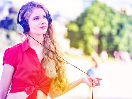 Girl in headphones and red shirt listens to mp3 music in city park. Teen portrait with hands to head backlight sun flare tone image.の写真素材