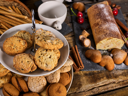 Oatmeal cookies and rolled cake on tier cake stand with cherry and nut on kitchen on wooden table in village style. Two cups of tea on wooden table. Perfect product.の写真素材