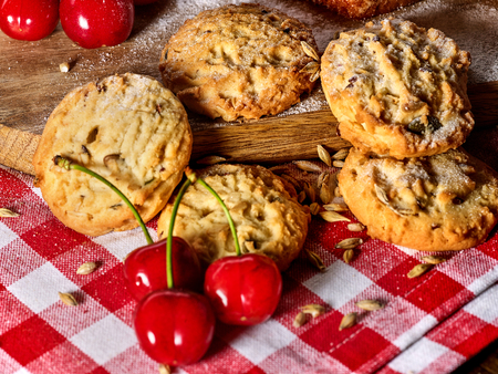 Oatmeal cookies with husk oats and cherry on kitchen cutting board gingham checkered cotton fabric on table in village style for picnic close up. Added sweet cherry. Culinary exhibition.の写真素材