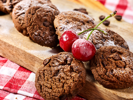 Oatmeal chocolate cookies with coffee grains and cherry, powdered sugar on kitchen cutting board with checkered fabric on wooden table in village style for picnic. Improved mood.の写真素材