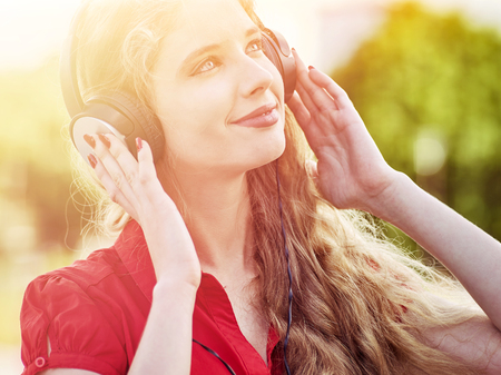 Girl in headphones and red shirt listens to mp3 music in city park. Teen portrait with hands to head backlight sun flare tone image. General cultural development of student.の写真素材