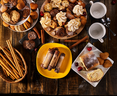 Meringue kisses, oatmeal cookies and sand chocolate cake on tier cake stand. Crispy wafer rolls with cream and cherry berry on cutting round board on wooden table. Perfect product.の写真素材