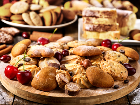 Oatmeal cookies and sand chocolate cake with cherry berry and crispy wafer rolls with cream on cutting board on wooden table in rustic style. Problems with excess weight. Sunlight in kitchen.の写真素材