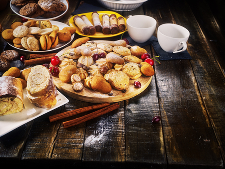 Oatmeal cookies and sand chocolate cake with cherry berry and crispy wafer rolls with cream on cutting board on wooden table in rustic style. Sunlight in kitchen.の写真素材