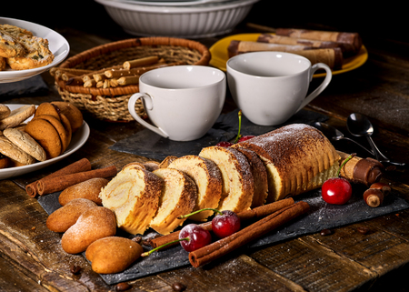 Sand cookies heart shape, rolled cake with cherry and cinnamon stick, coffee cups on kitchen on wooden table in village style. Useful for digestion. Perfect product.の写真素材