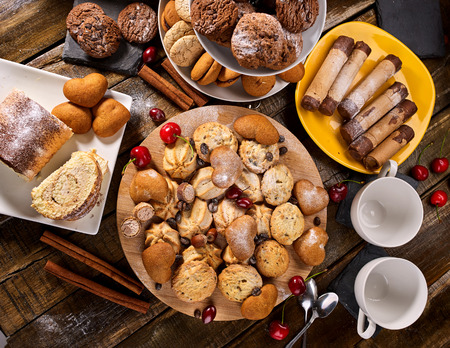 Oatmeal cookies and sand chocolate cake with cherry berry and crispy wafer rolls with cream on cutting board on wooden table in rustic style. Factory for production of sweets.の写真素材