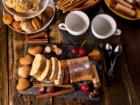 Oatmeal and nut cookies heart shape and sand rolled biscuit on tier cake stand and cinnamon stick on kitchen on wood table in rustic style. Cups of tea and dessert spoon. Top view.の写真素材