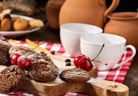 Oatmeal chocolate cookies with coffee grains and cherry, powdered sugar on kitchen cutting board with checkered fabric on wooden table in village style for picnic. Breakfast for loved one.の写真素材