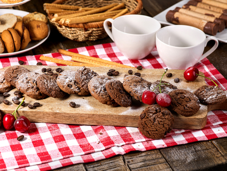 Oatmeal chocolate cookies with coffee grains and cherry, powdered sugar on kitchen cutting board with checkered fabric on wooden table in village style for picnic. Delight pleasant taste.の写真素材