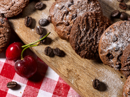 Oatmeal chocolate cookies with coffee grains and cherry, powdered sugar on kitchen cutting board with checkered fabric on wooden table in village style for picnic. Supplement for breakfast.の写真素材