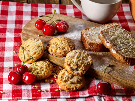 Oatmeal cookies with husk oats and cherry cupcake with powdered sugar on kitchen cutting board and cup of tea gingham checkered cotton fabric on table in village style for picnic. Coffee in cafe.の写真素材