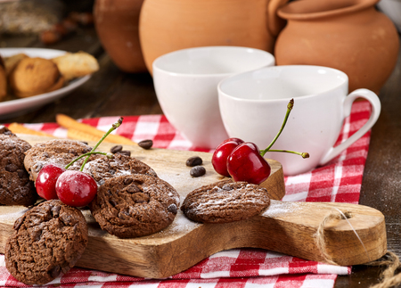 Oatmeal chocolate cookies with coffee grains and cherry, powdered sugar on kitchen cutting board with checkered fabric in village style for picnic. Macaron in background. It has pleasant taste.の写真素材