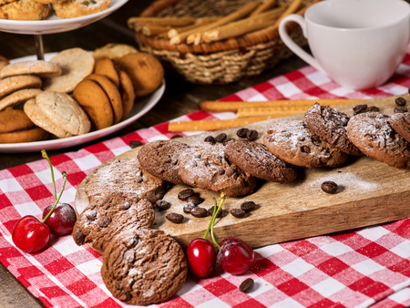 Oatmeal chocolate cookies with coffee grains and cherry, powdered sugar on kitchen cutting board with checkered fabric on wooden table in village style for picnic. Breakfast for loved one.の写真素材