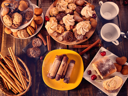 Meringue kisses, oatmeal cookies and sand chocolate cake on tier cake stand. Crispy wafer rolls with cream and cherry berry on cutting round board on wooden table in rustic style top view.の写真素材