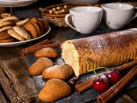 Oatmeal cookies and rolled cake on tier cake stand with cherry and nut on kitchen on wooden table in village style. Two cups of tea on wooden table. Rejection of many products.の写真素材