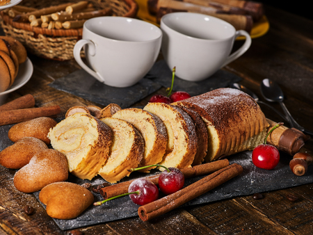 Sand cookies heart shape , rolled cake with cherry and cinnamon stick, coffee cups on kitchen on wooden table in village style.の写真素材