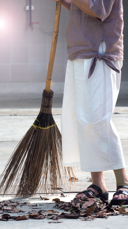 Old woman is sweeping dry leaf on the outdoor cement floor by big long broom.の写真素材