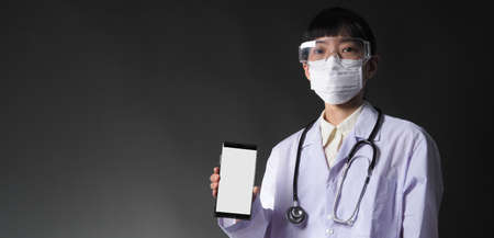 Middle aged of asian woman doctor showing mobile phone screen to communicate something. And she wearing white lab suit, mask and goggle to prevent coronavirus or covid-19の写真素材
