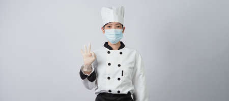 Asian woman chef in white color uniform with hygienic such as medical face mask and rubber hand glove for prevent or protect covid-19 or coronavirus pandemic. Studio shot and copy space.の写真素材