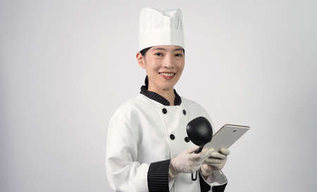 Asian woman chef holding smartphone or digital tablet and received food order from online shop or merchant application. she smiling in chef uniform and standing in studio with white color background.の写真素材