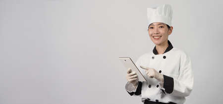 Asian woman chef holding smartphone or digital tablet and received food order from online shop or merchant application. she smiling in chef uniform and standing in studio with white color background.の写真素材