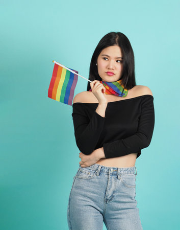 Portrait of young girl holding an LGBT flag standing against a blue green background studio. Asian LGBTQ woman with rainbow scarf on neck. look smart bright and energetic cheerful. LGBTQ concept.の写真素材