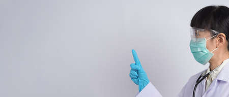 Doctor in uniform points to an empty area. Wearing blue medical latex gloves and clear goggles and green N95 mask to prevent epidemic virus. Asian doctor. Hand gesture. Medical stethoscope. Virus bookの写真素材