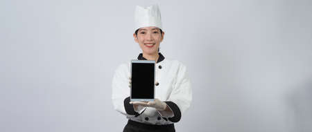 Asian woman chef holding smartphone or digital tablet and received order from online shop or merchant application. she smiling in chef uniform and standing in white background. Online food merchant.の写真素材