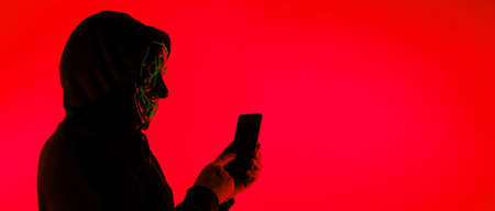 Anonymous hacker and face mask with smartphone in hand. Man in black hood shirt trying to hack...