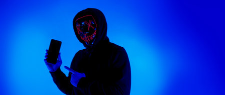 Anonymous hacker and face mask with smartphone in hand. Man in black hood shirt holding and using mobile phone on white background. Represent cyber crime data hacking or stealing personal data conceptの写真素材