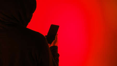 Anonymous hacker and face mask with smartphone in hand. Man in black hood shirt holding and using mobile phone on white background. Represent cyber crime data hacking or stealing personal data conceptの写真素材
