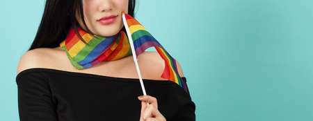 LGBTQ woman holding pride flag standing against a blue green background. Asian LGBTQ woman with rainbow scarf on neck. look smart bright and energetic cheerful. LGBTQ diversity pride concept.の写真素材