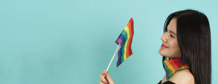 LGBTQ woman holding pride flag standing against a blue green background. Asian LGBTQ woman with rainbow scarf on neck. look smart bright and energetic cheerful. LGBTQ diversity pride concept.の写真素材