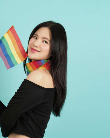 LGBTQ woman holding pride flag standing against a blue green background. Asian LGBTQ woman with rainbow scarf on neck. look smart bright and energetic cheerful. LGBTQ diversity pride concept.の写真素材