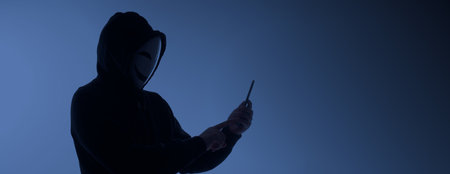 Hacker Anonymous and face mask with smartphone in hand. Man in black hood shirt holding and using mobile phone on white background. Represent cyber crime data hacking or stealing personal data conceptの写真素材