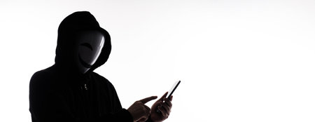 Hacker Anonymous and face mask with smartphone in hand. Man in black hood shirt holding and using mobile phone on white background. Represent cyber crime data hacking or stealing personal data conceptの写真素材