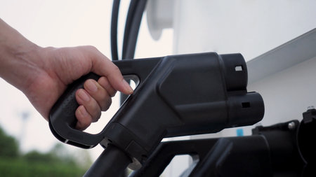 Hand holding EV charging plug in front of camera with blurred background of outdoor. Driver of electric car hold batteries charger plug. Sustainable energy powered electric charging station backgroundの写真素材
