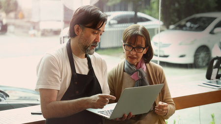 A friendly middle aged barista and a senior businesswoman collaborate using a smartphone and laptop inside a modern cafe. They appear to discuss online orders digital marketing or business strategy.の写真素材