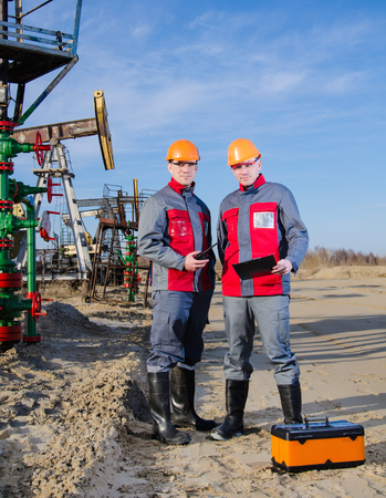 Workers in the oilfield, one holding radio in his hand. Pumpjack and wellhead background. Toolbox foreground. Oil and gas concept.の写真素材