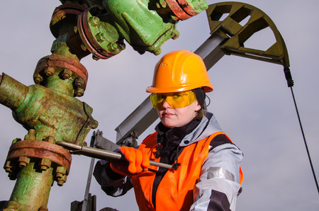 Woman engineer in the oil field repairing wellhead with the wrench wearing orange helmet and work clothes. Pump jack background. Oil and gas concept.の写真素材