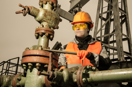 Woman engineer in the oil field repairing wellhead with the wrench wearing orange helmet and work clothes. Oil and gas concept. Toned.の写真素材