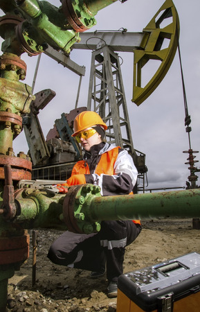 Woman engineer in the oil field repairing wellhead with the wrench wearing orange helmet and work clothes. Pump jack and tool box background. Oil and gas concept.の写真素材