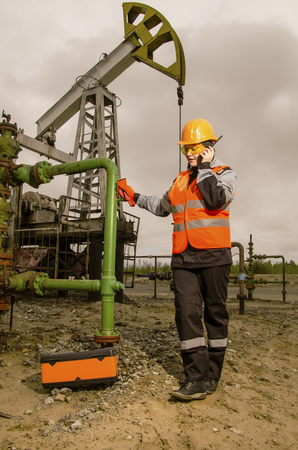 Woman engineer in the oil field talking on the radio wearing orange helmet and work clothes. Pump jack background. Oil and gas concept. Toned.の写真素材
