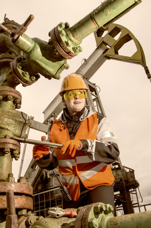 Woman engineer in the oil field repairing wellhead with the wrench wearing orange helmet and work clothes. Pump jack background. Oil and gas concept. Toned.の写真素材